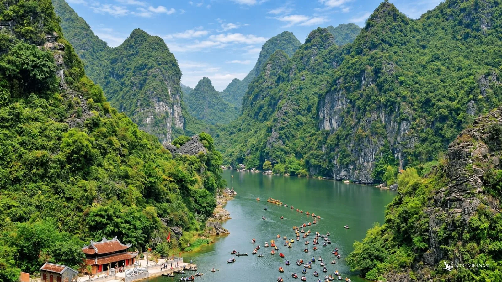 A rowboat passing a riverside temple beneath an overhanging limestone cliff