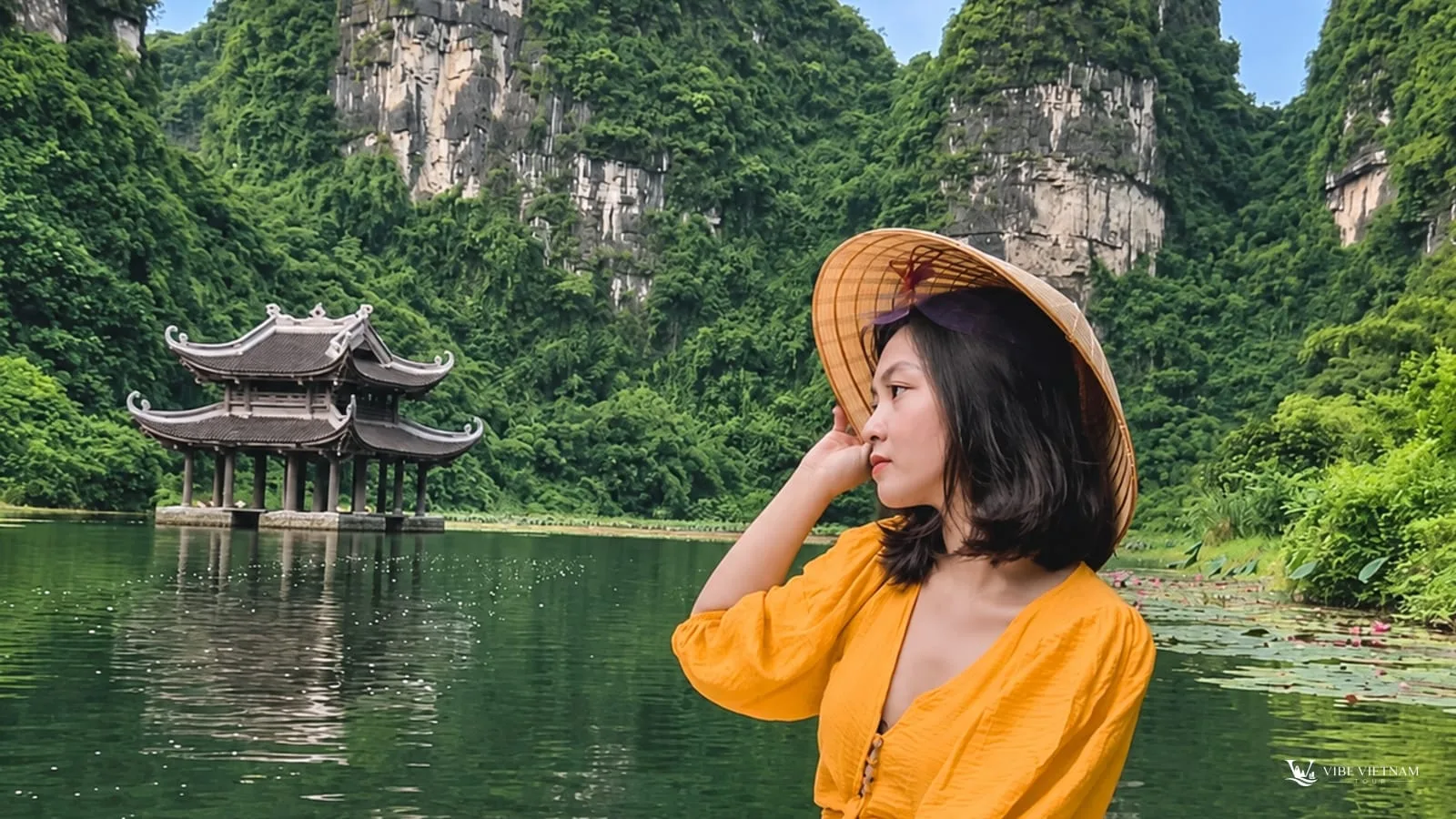 A visitor in a traditional conical hat beside the water, with a pavilion and karst peaks behind her